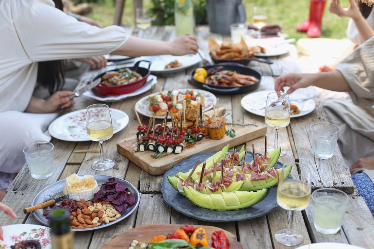 A long picnic table outdoors, set with shared plates and wine